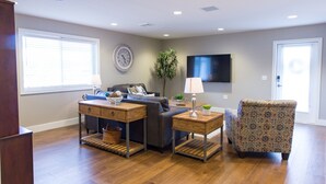 A living room with a sofa, two side tables, a patterned armchair, a clock, a television, and a window with blinds. - Downtown Getaway (Caseville)
