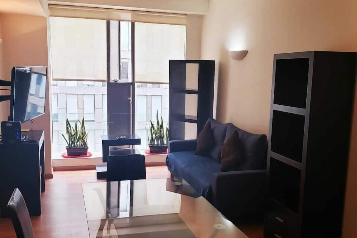 A modern living room with a glass coffee table, a sofa, and a TV on a stand. There are two potted plants and a bookshelf.