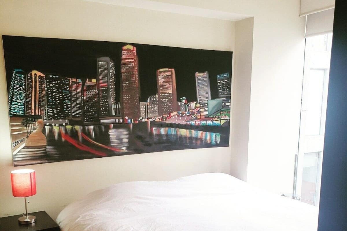 A hotel room with a large cityscape painting on the wall, a bed with white linens, and a bedside table with a lamp.