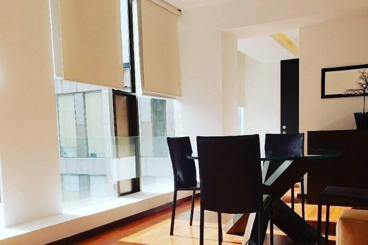 A modern living room with a wooden floor, a sofa, a dining table with chairs, and a large window with blinds.