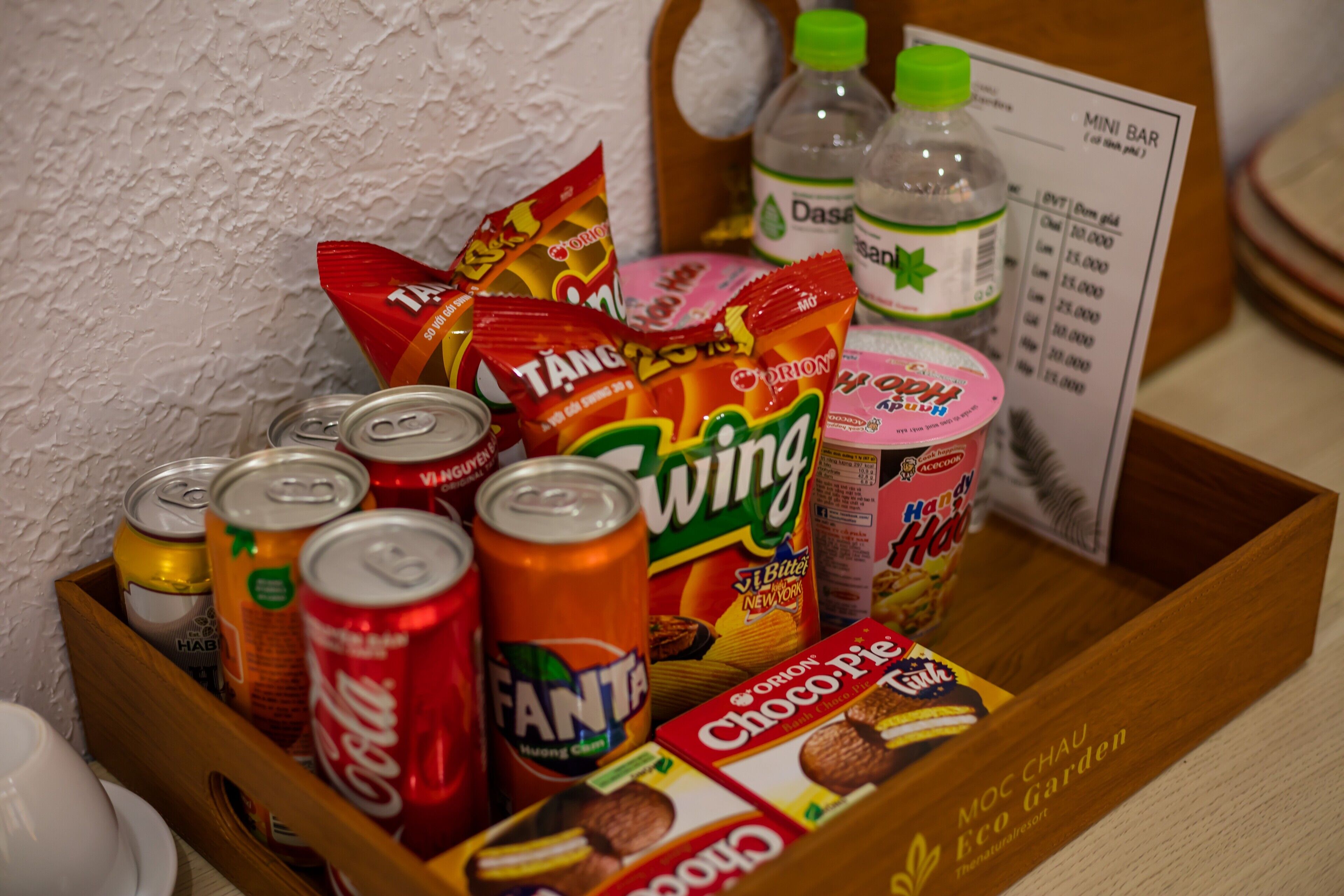 A wooden tray with various snacks and drinks, including cans of Fanta and Coca-Cola, a pack of Tang, a bottle of Dasani water, and a box of Choco Pie.