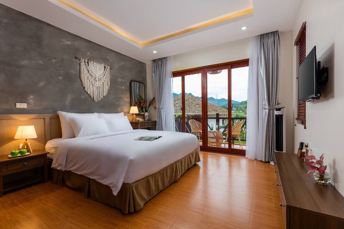 Ein Hotelzimmer mit einem großen Bett, einem Fernseher, einem Balkon mit Blick und einem an der Wand montierten Fernseher.