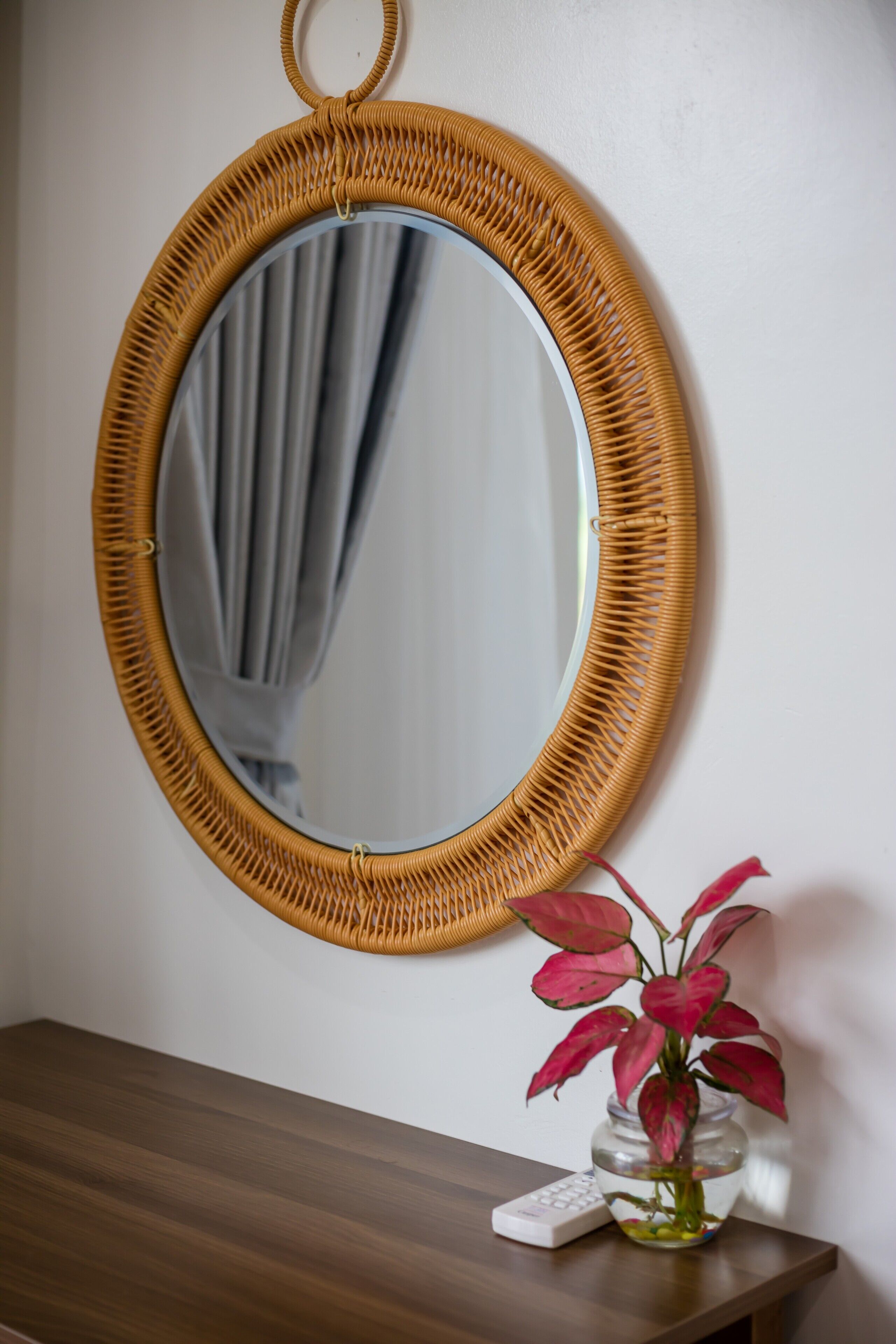 A round mirror with a woven frame, a vase with pink flowers, and a remote control on a wooden surface.