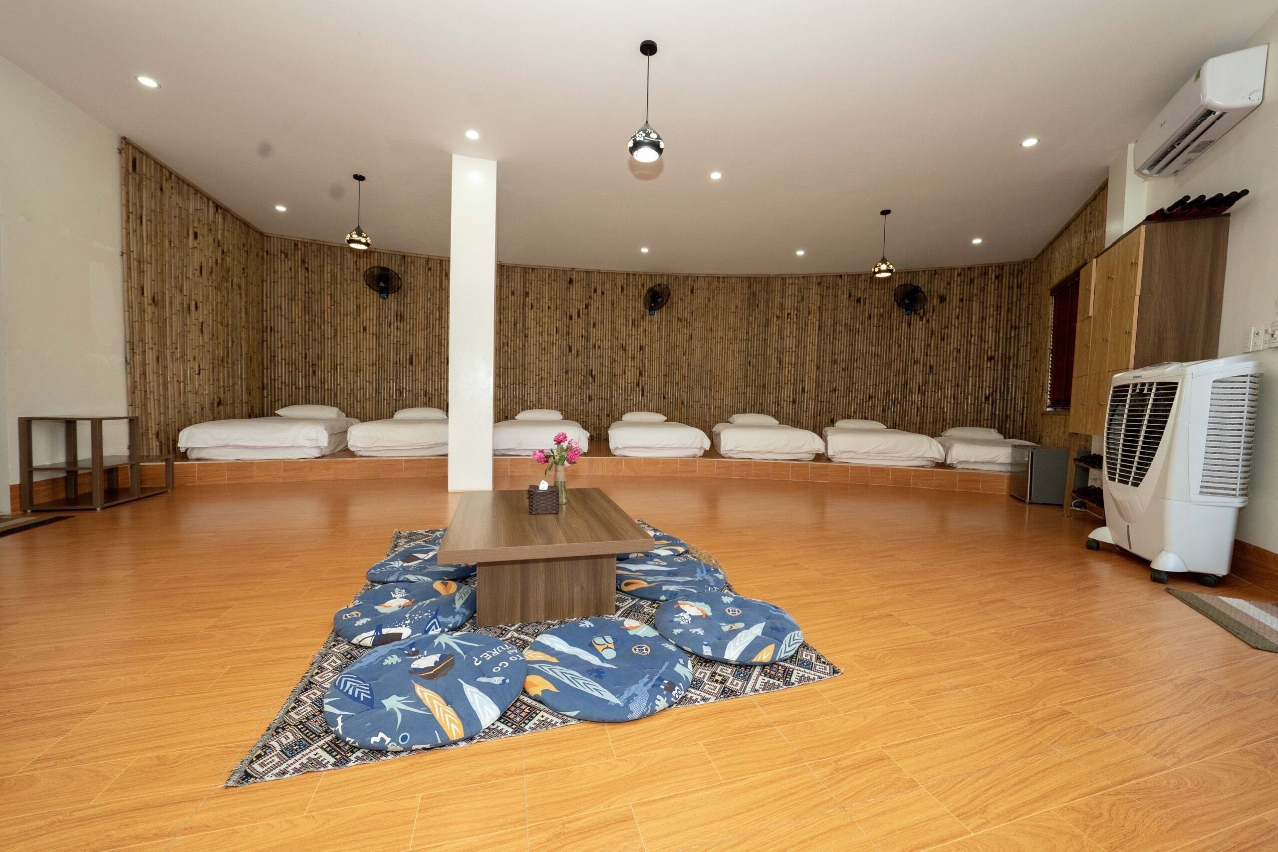 Amplia habitación con suelo de madera, una mesa central y varias zonas de estar con cojines blancos.
