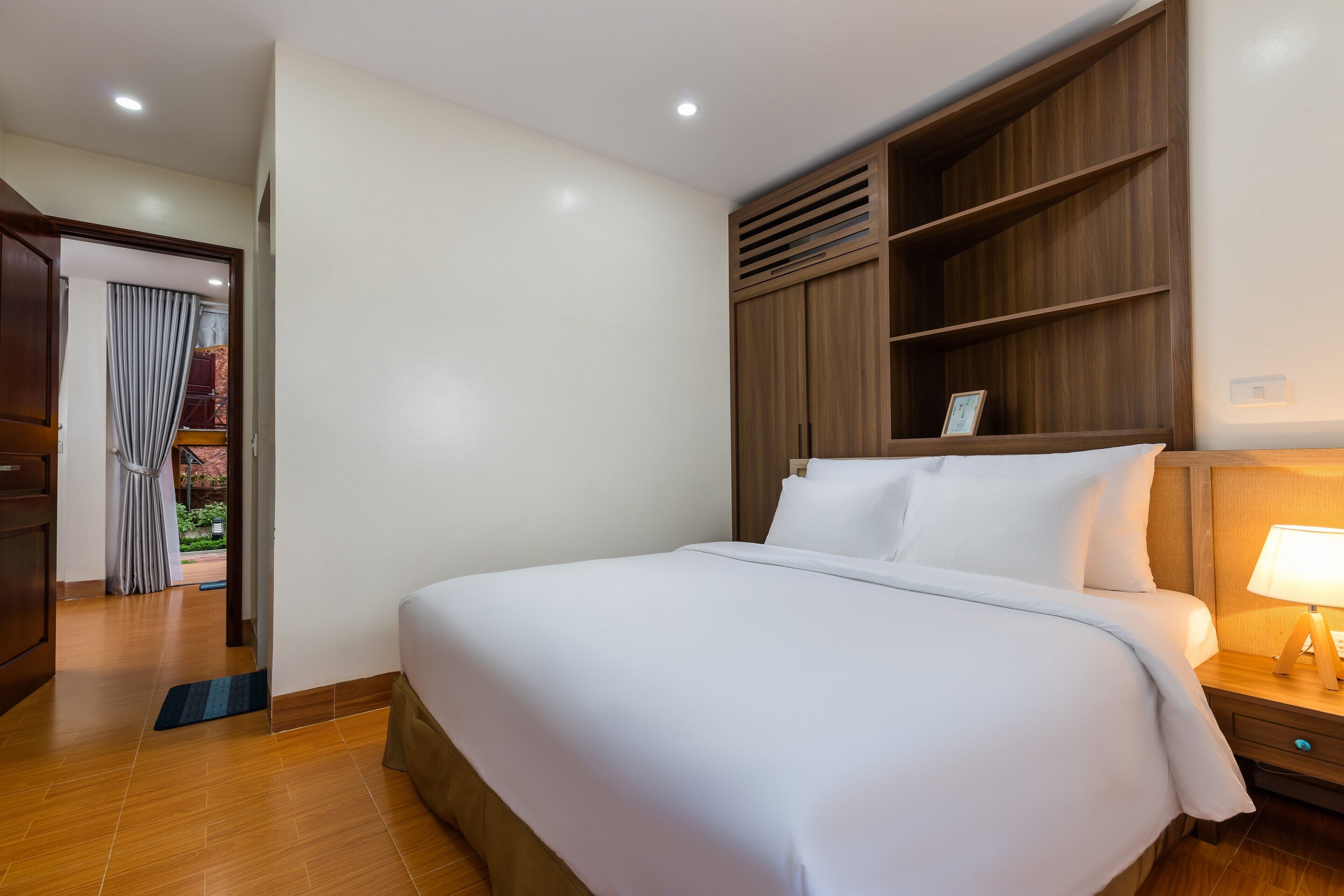 Habitación de hotel con una cama grande, cabecero de madera, estantes empotrados, una mesita de noche con lámpara y vistas a un balcón a través de una puerta con cortinas.