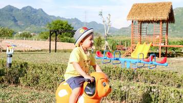 A child riding a yellow elephant-shaped toy on a grassy field with a playground and mountains in the background.