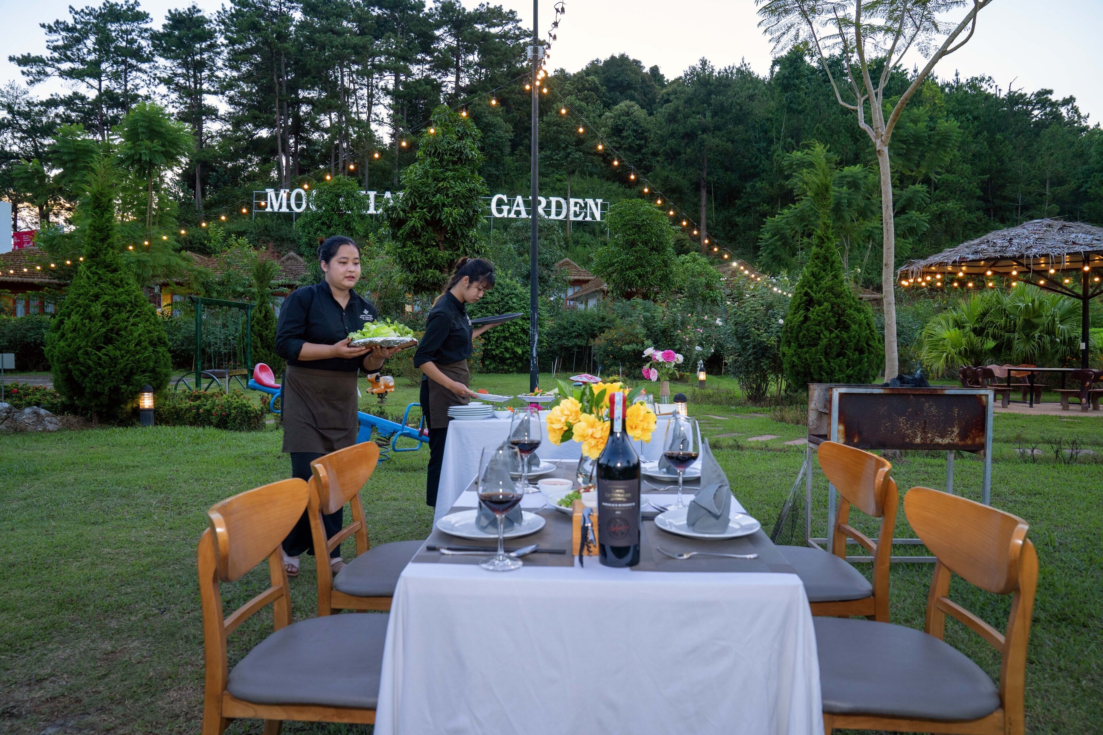 An outdoor dining area with tables set for a meal, surrounded by greenery and string lights.
