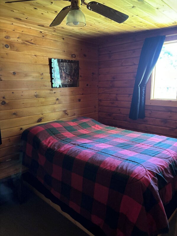 A wooden cabin room with a bed, a ceiling fan, a window with curtains, and a wall-mounted picture.
