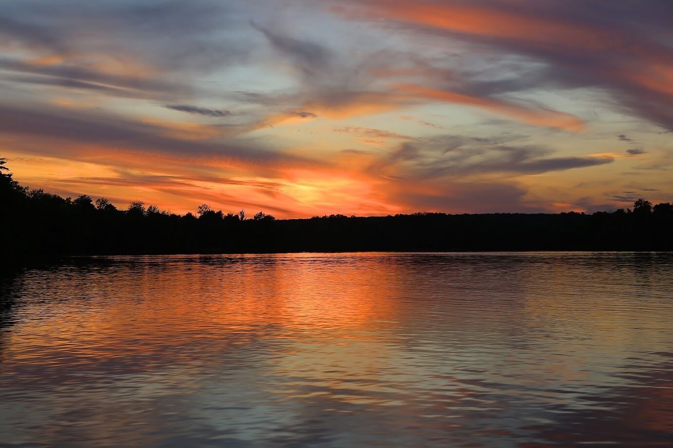 A sunset over a calm lake with a silhouette of trees in the background.