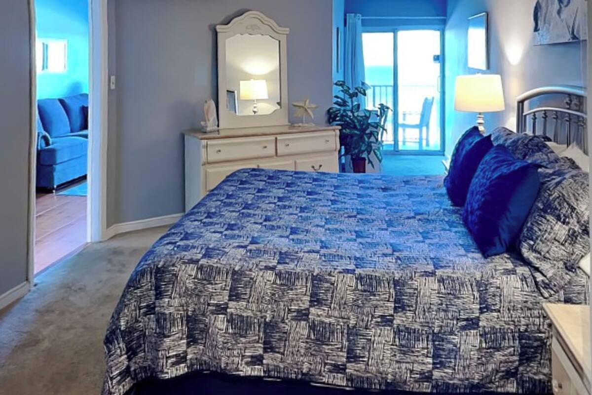 A bedroom with a large bed, a dresser, a mirror, and a view of the outside.
