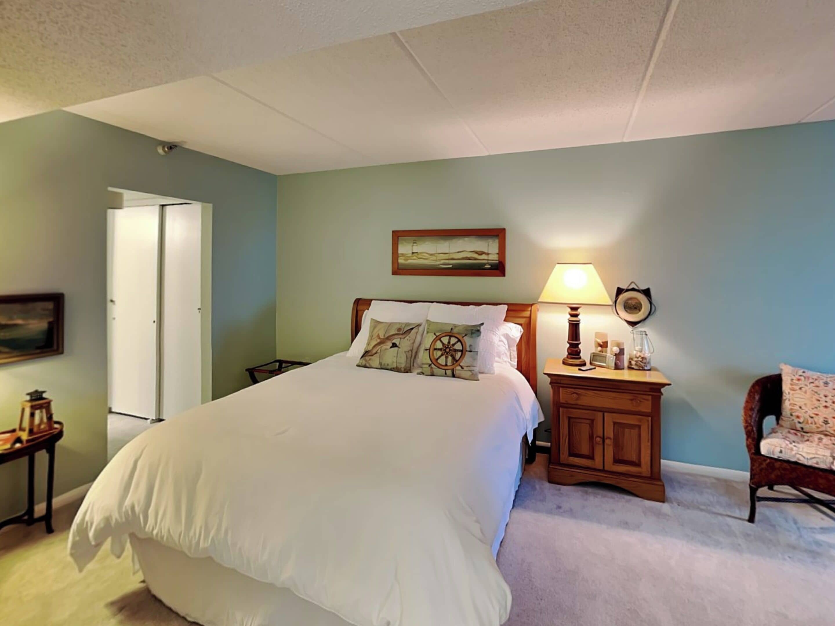 A bedroom with a large bed, a nightstand with a lamp, a chair, and a door.