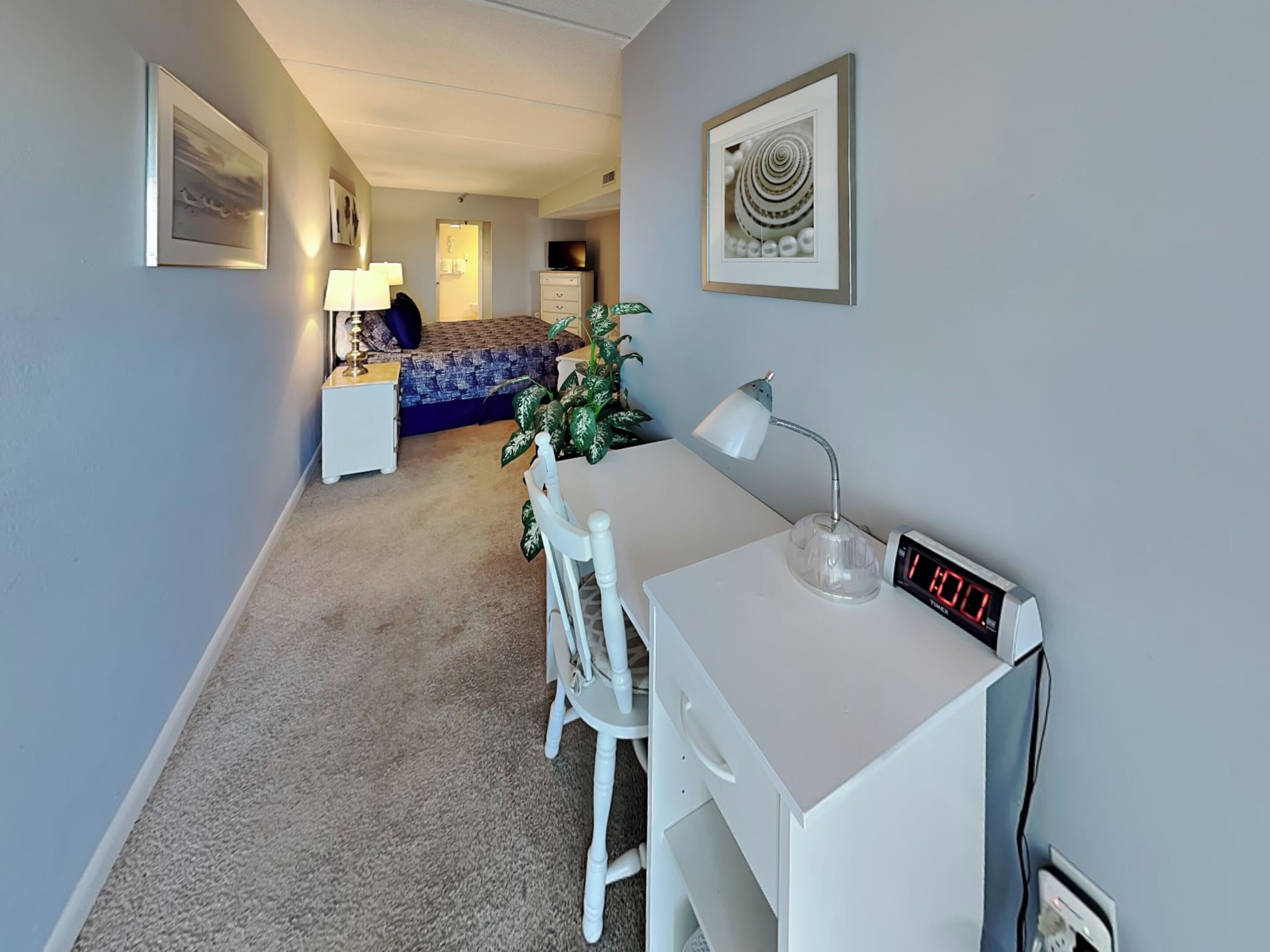A hotel room with a desk, chair, lamp, clock, and a bed with a blue and white patterned bedspread.