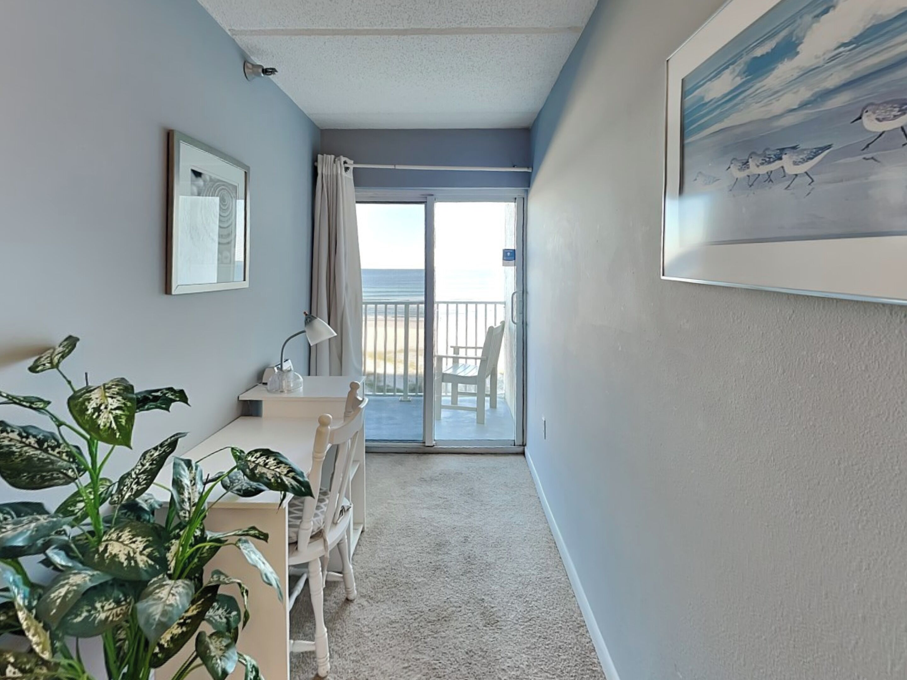 A room with a desk, chair, and a potted plant. There is a door leading to a balcony with a view of the beach.