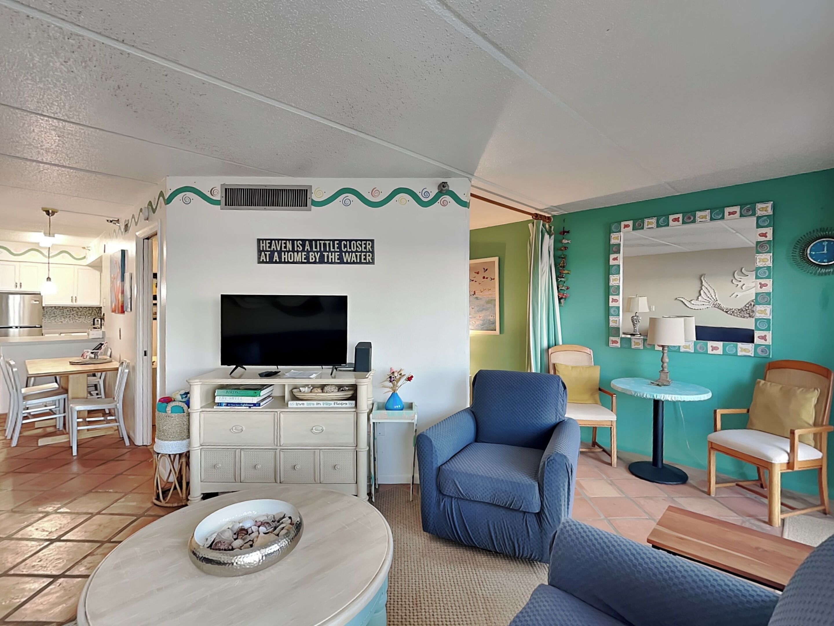 Un salon avec un téléviseur, un fauteuil bleu, une petite table avec un saladier, et un coin cuisine visible à travers une porte ouverte.