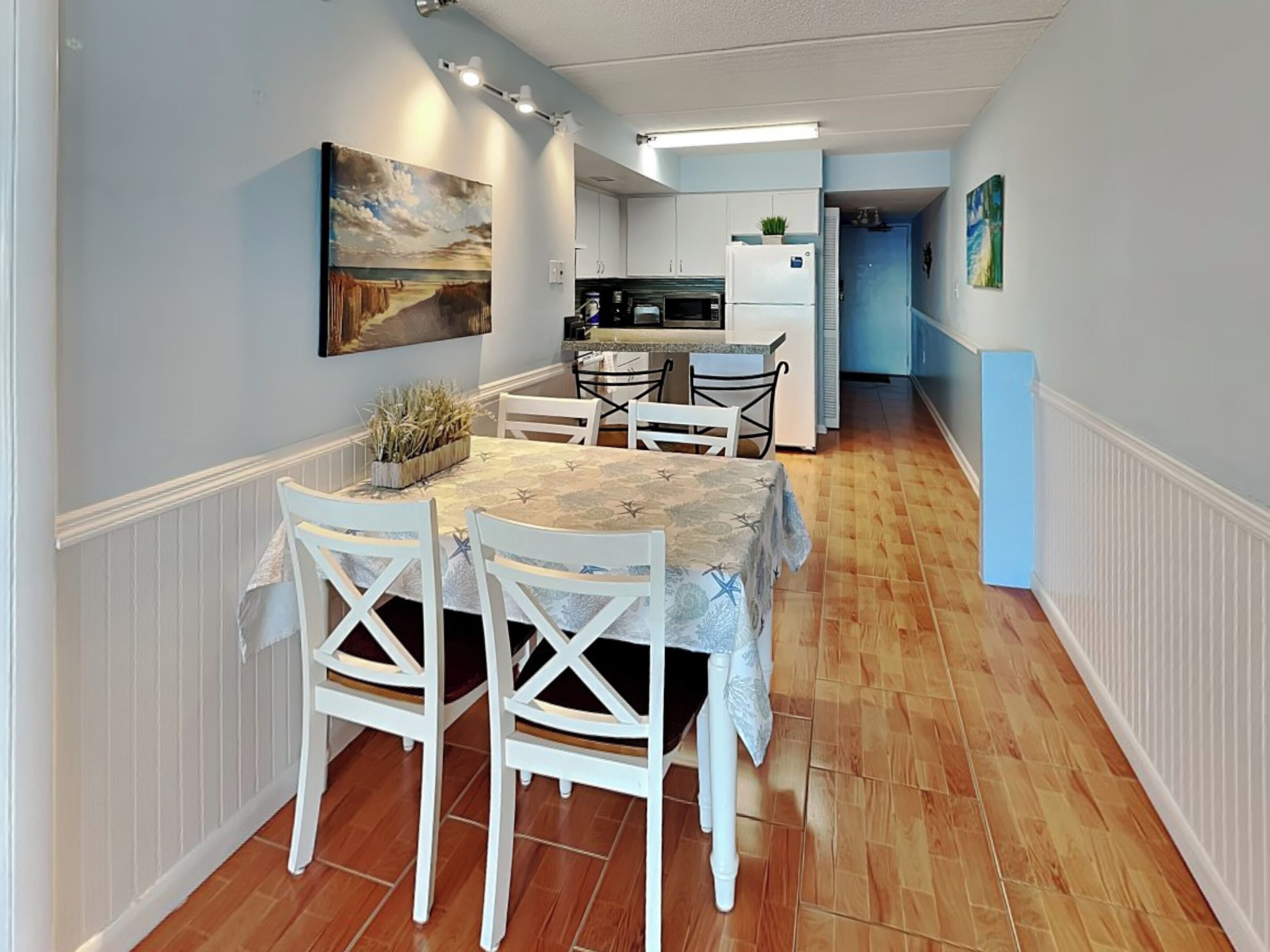 Un coin repas avec une table dressée pour quatre personnes, des chaises blanches, un tableau au mur et une cuisine en arrière-plan.