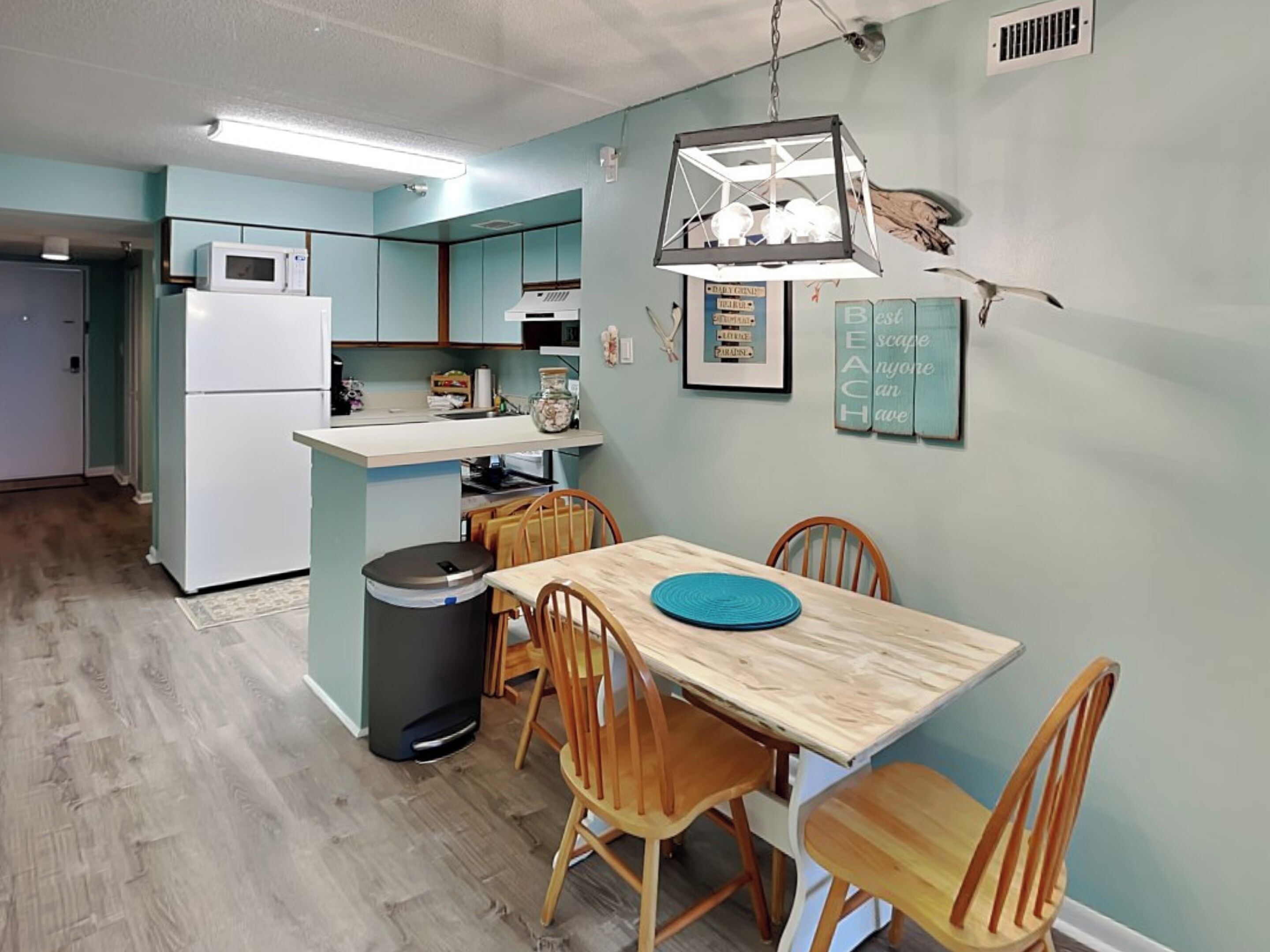 A compact kitchen with a refrigerator, microwave, and a small island. There is a dining table with chairs and a trash bin.