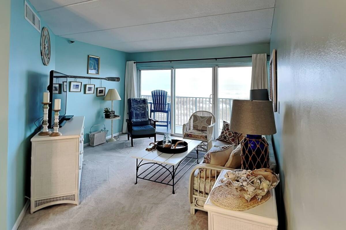 A room with a large sliding glass door leading to a balcony, a white dresser, a blue armchair, and a wicker chair.