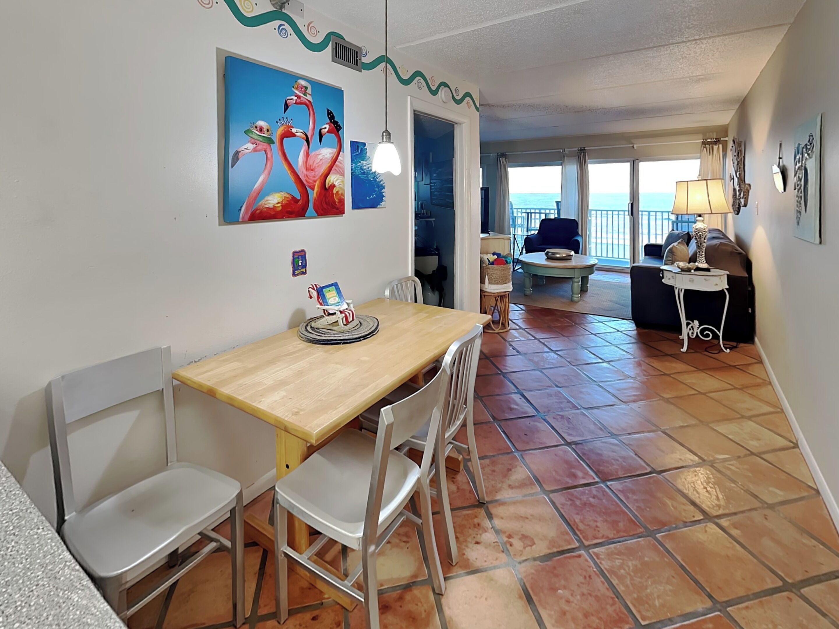 Een eetgedeelte met een houten tafel en witte stoelen, een schilderij van flamingo's, en uitzicht op zee door grote ramen.