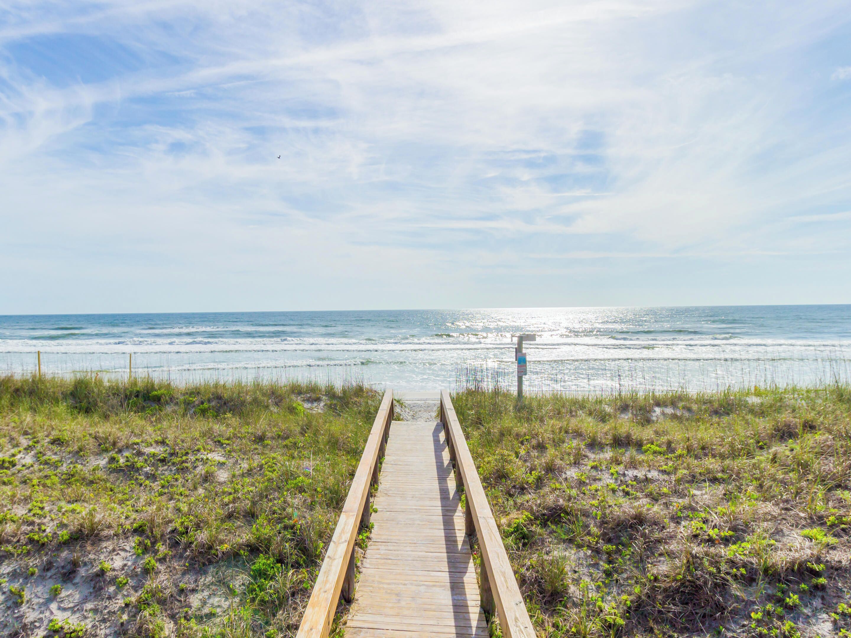 A wooden boardwalk leading to the beach with a clear view of the ocean and a signpost in the distance.