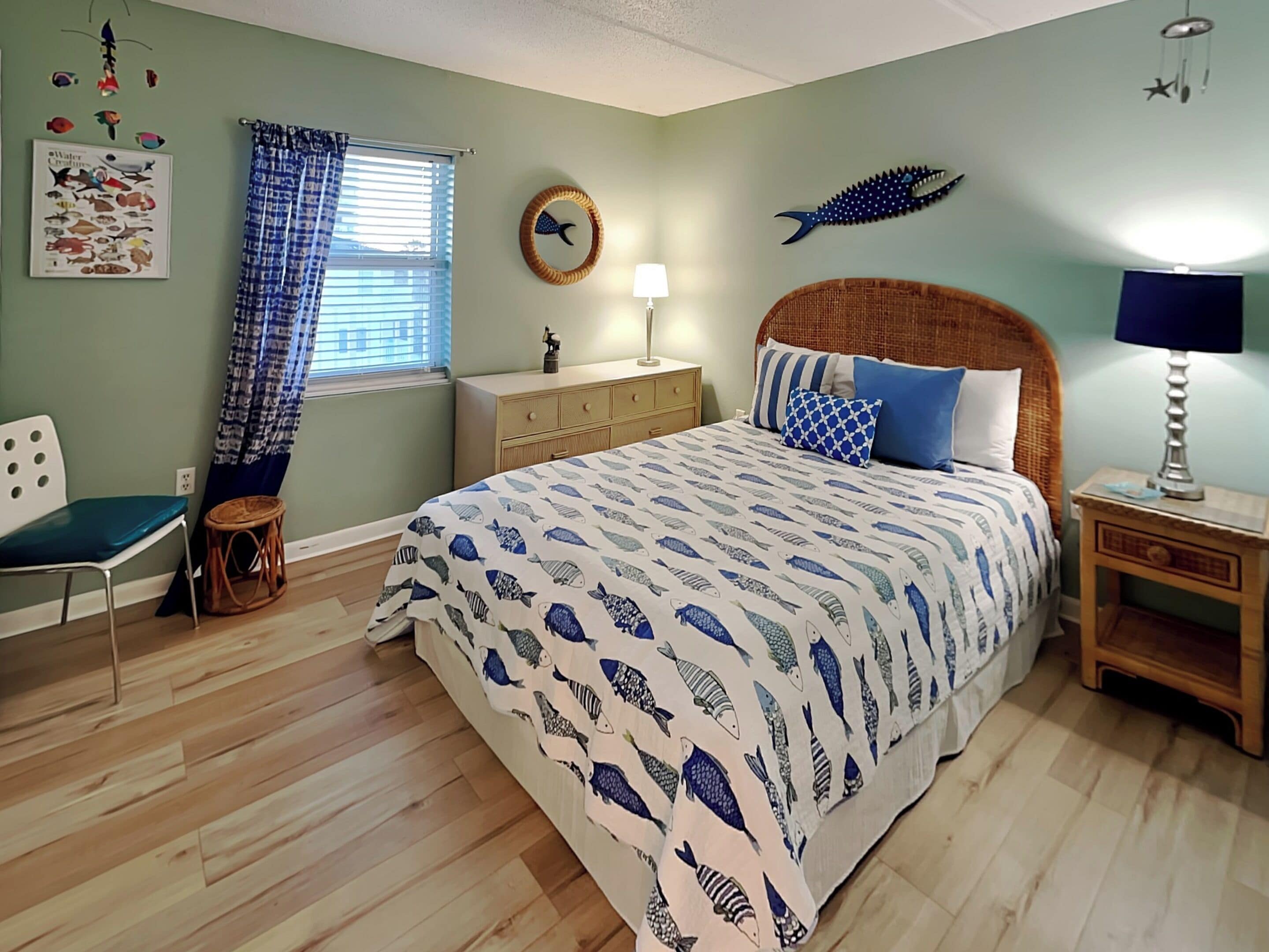 Een slaapkamer met een bed, een stoel, een nachtkastje, een lamp, een dressoir, een raam met gordijnen, en wanddecoraties waaronder een vis en een poster.