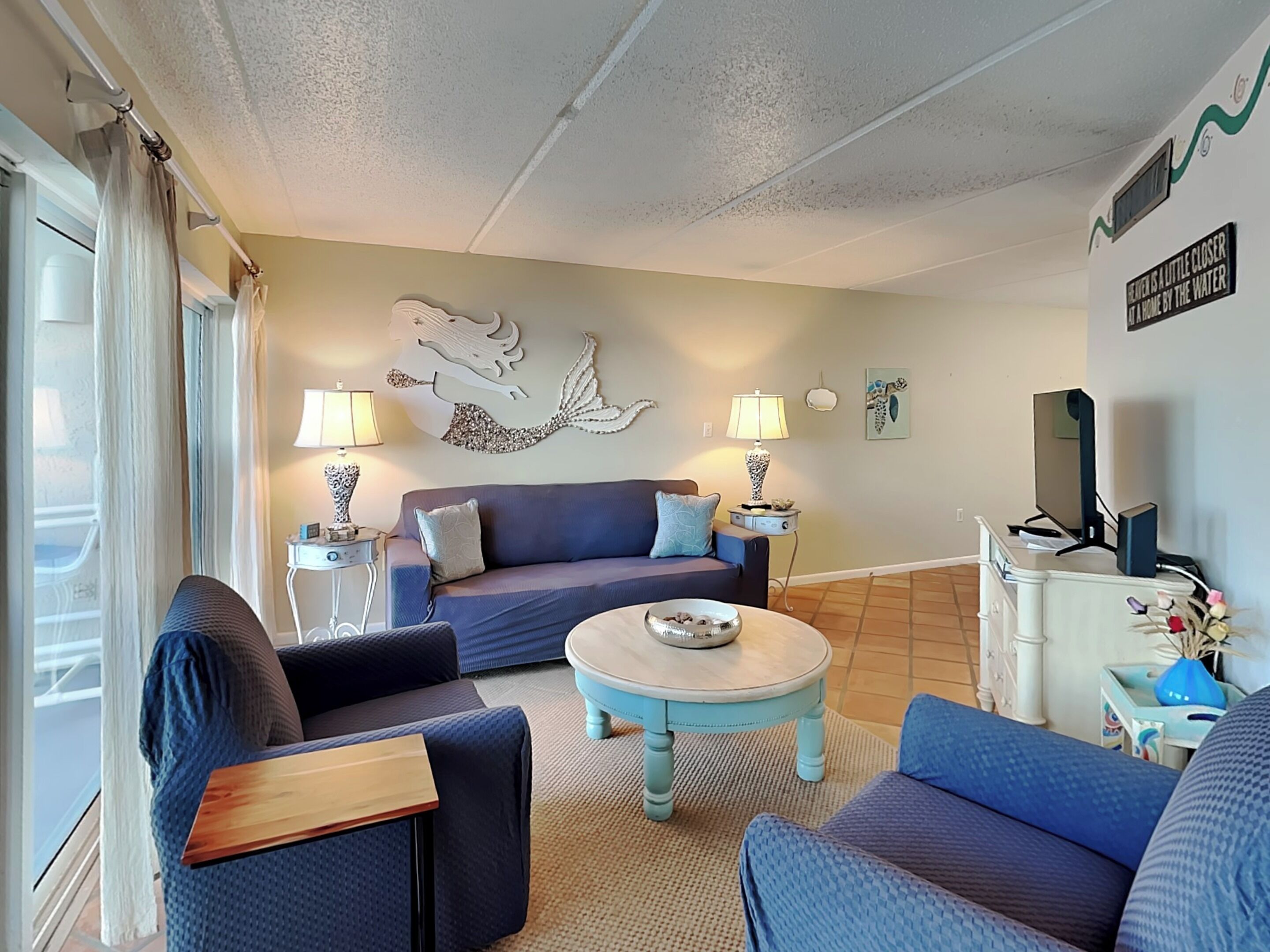 A living room with a sofa, two armchairs, a coffee table, and a TV. There are two lamps on side tables, and a decorative wall piece above the sofa.