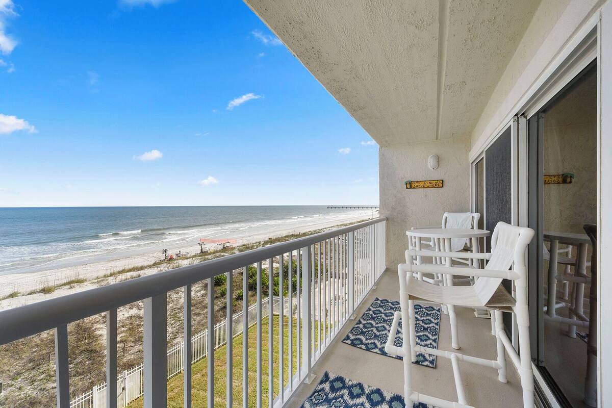 A balcony with a view of the beach and ocean, white chairs, and a small table.