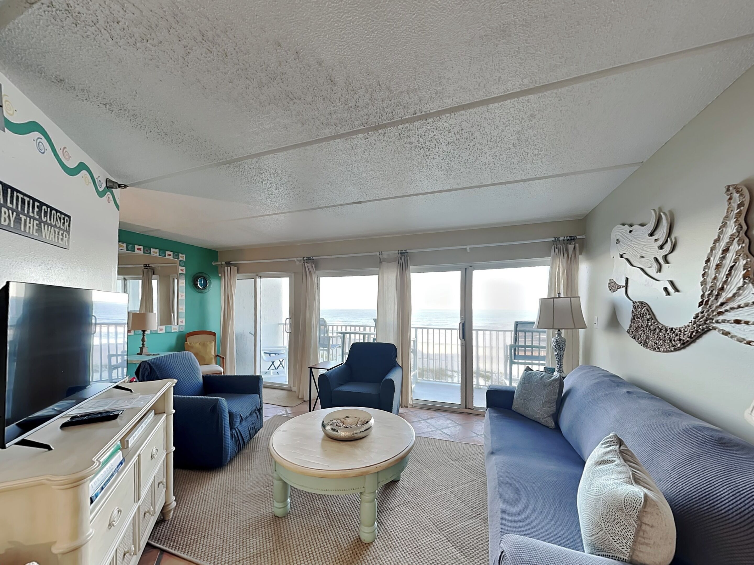 A spacious living room with a large TV, comfortable seating, a balcony with a view of the sea, and a decorative wall piece.