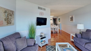 A living room with two grey sofas, a coffee table, a TV mounted on the wall, and a dining area in the background. - Jacksonville Beachdrifter by Distinctive Beach Rentals (Jacksonville Beach)