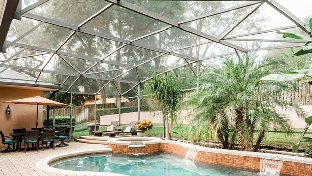 Una piscina interna con soffitto in vetro, circondata da palme e un patio con sedie e tavolo.