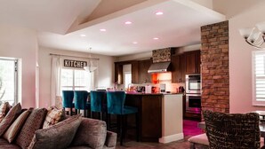 Una cucina moderna con una parete decorata in pietra, una zona pranzo con sedie color petrolio e un soggiorno con divano e cuscini fantasia.