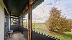 En balkong med utsikt över en gräsklädd äng och träd, en trästol och ett klinkergolv.