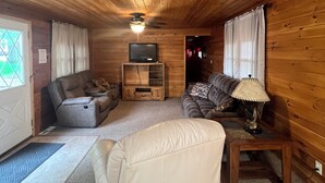 A cozy cabin interior with a wooden ceiling, a sofa, a recliner, a TV stand, and a lamp.