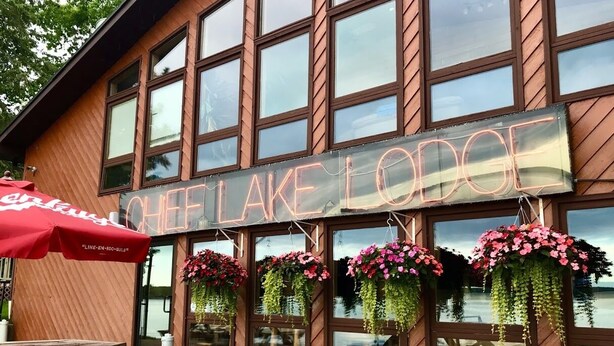 A building with a sign reading "CHEESE LAKE LODGE" and hanging flower baskets.