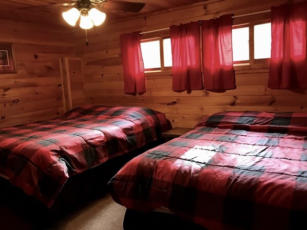 A cabin room with two beds, wooden walls, a ceiling fan, and a window with red curtains.