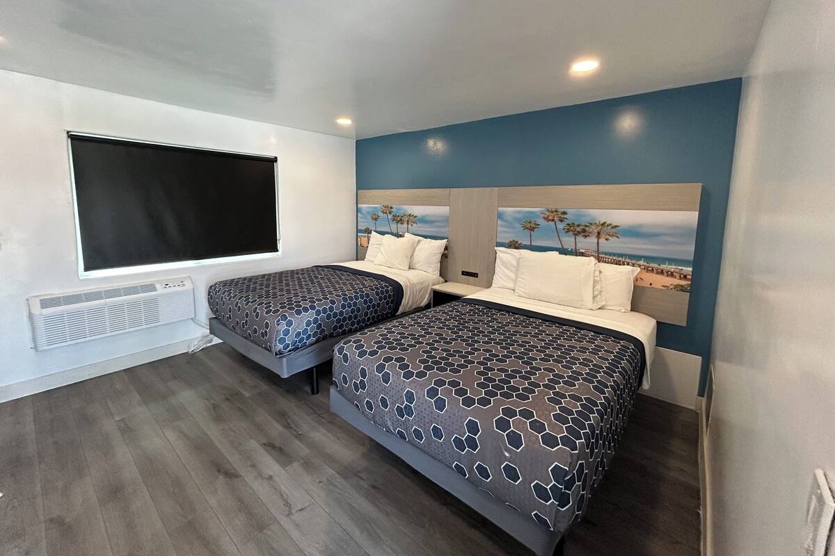 A room with two beds, a large window, a wall-mounted air conditioner, and a beach-themed mural on the wall.