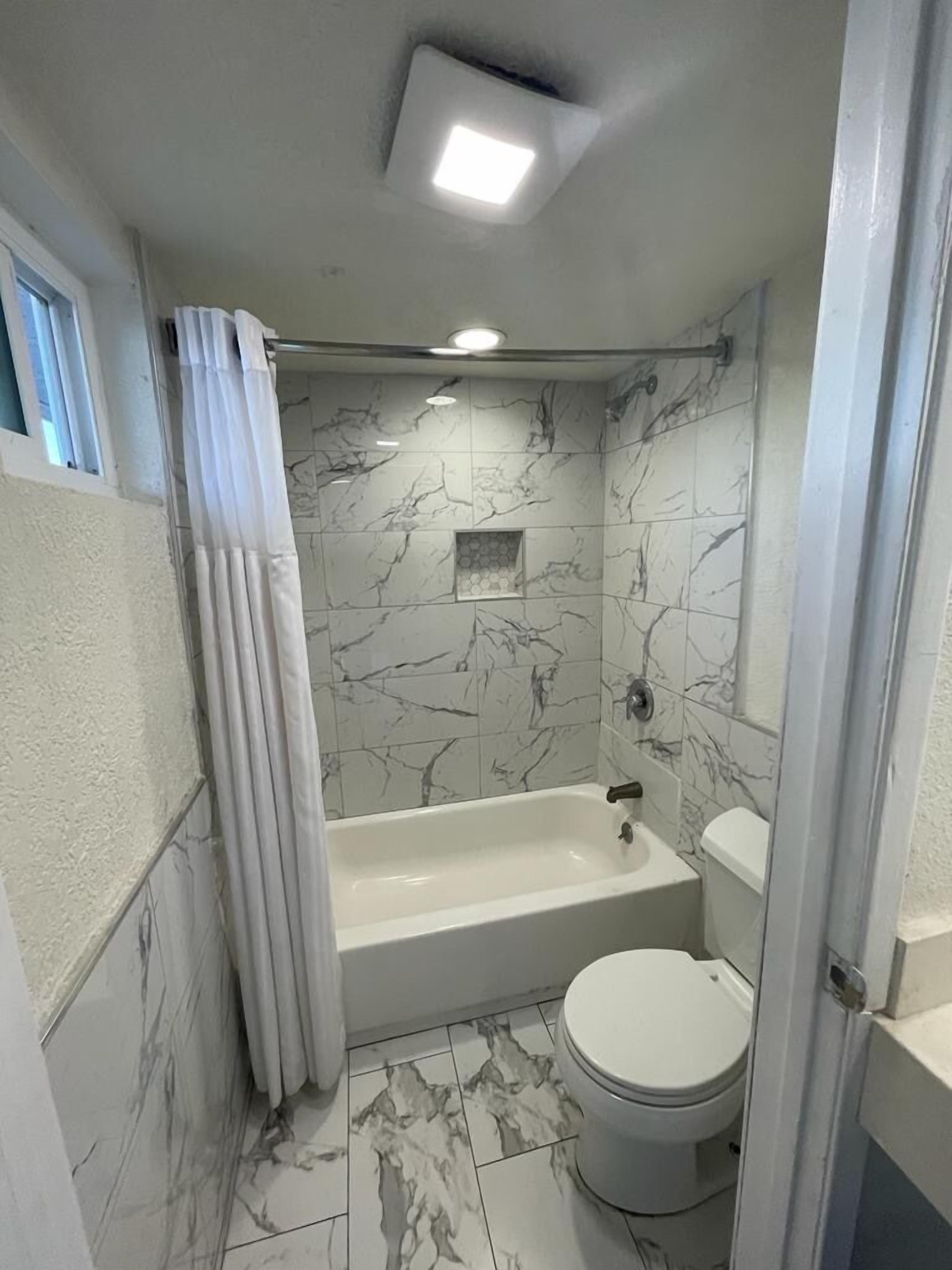A bathroom with a bathtub, toilet, and shower area, featuring marble tiles and a ceiling light.