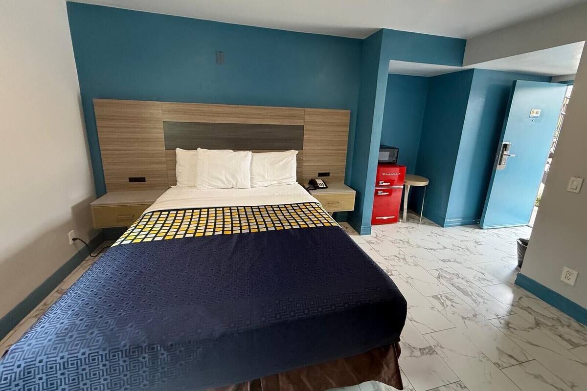 A hotel room with a bed, a nightstand, a red storage unit, and a door leading to another room.