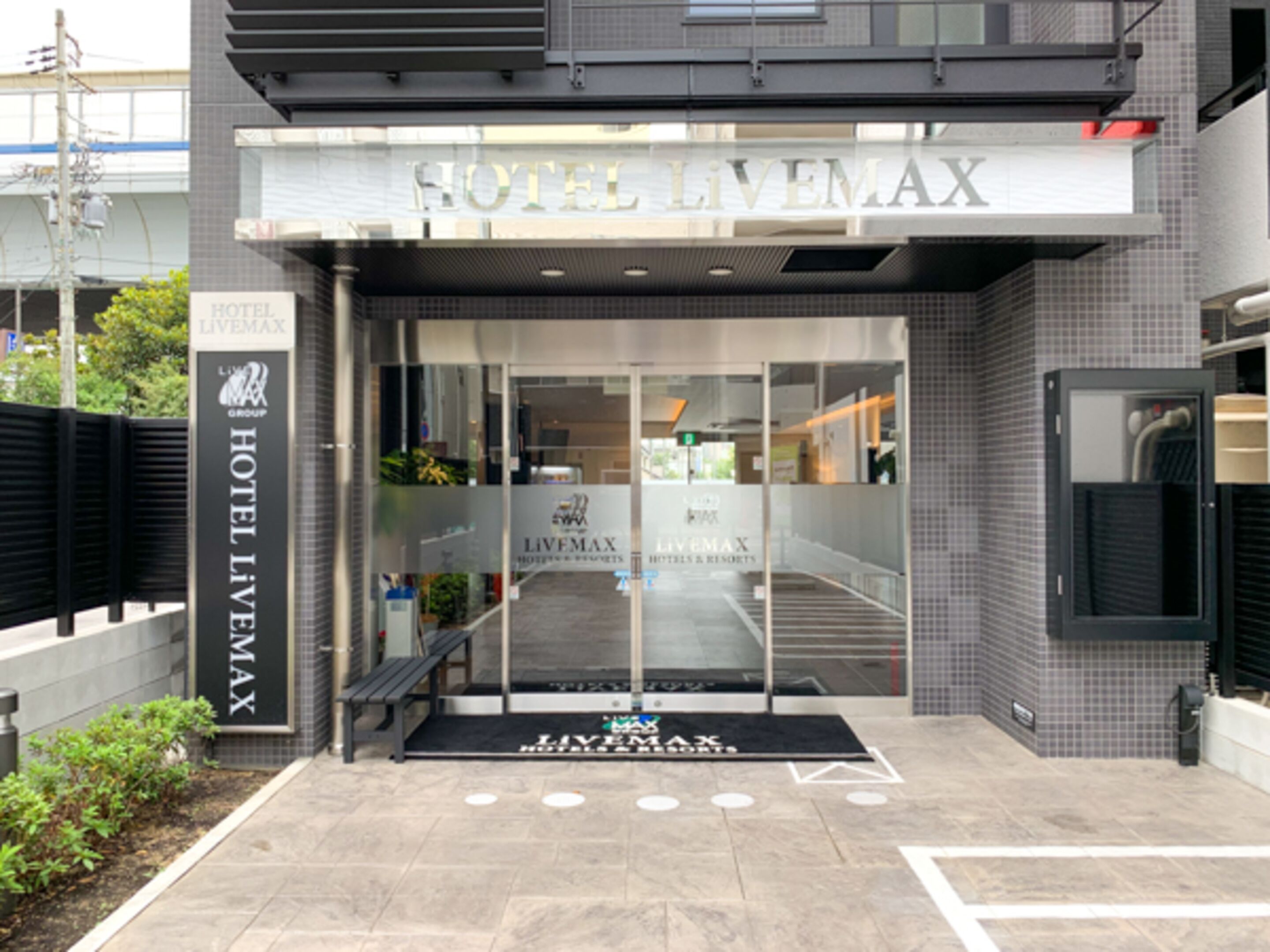 Der Eingang des Hotels Livemax mit modernem Design, inklusive Glastür und einer schwarzen Bank davor.