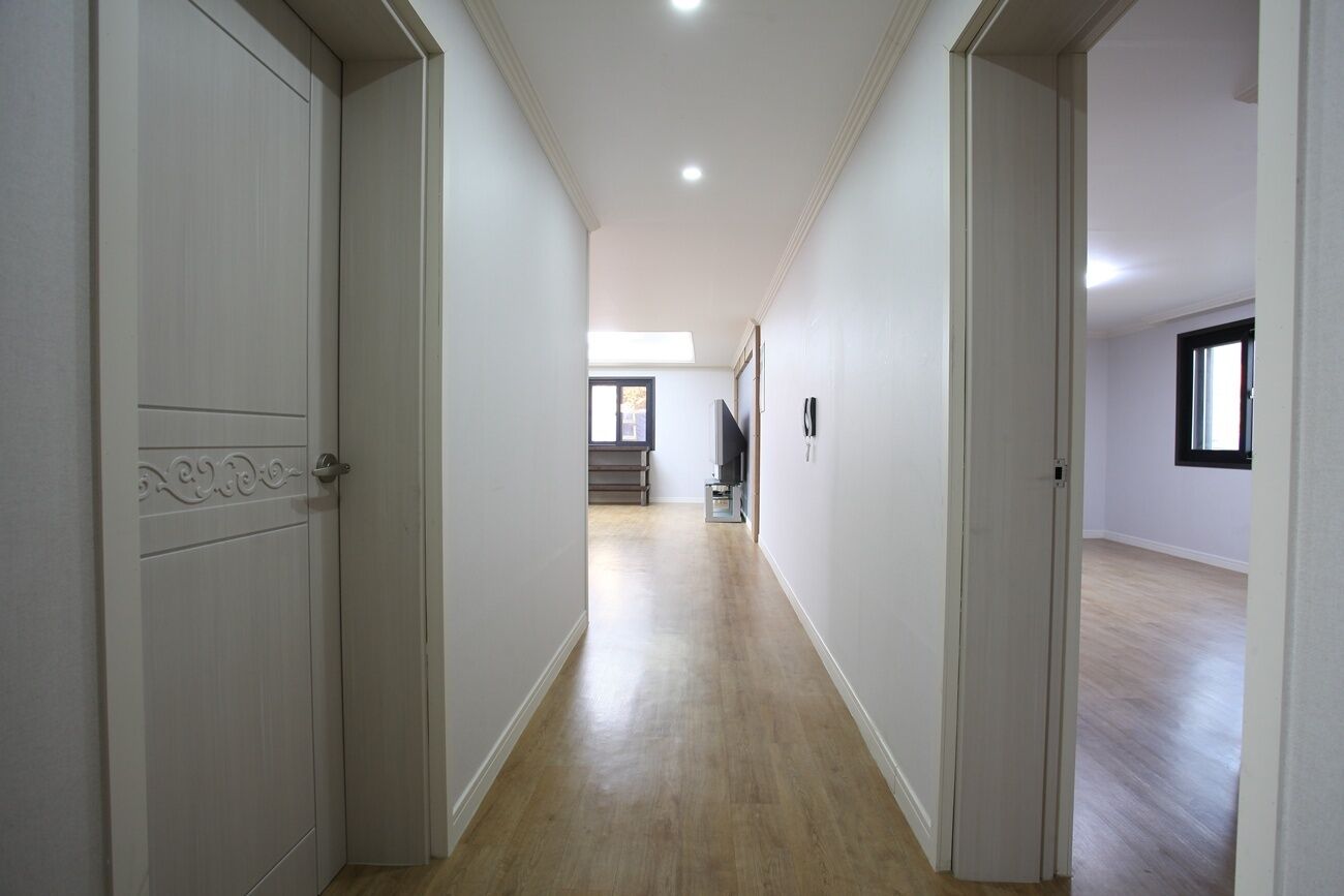 A hallway with white walls and wooden flooring, leading to a staircase and a room with a window.