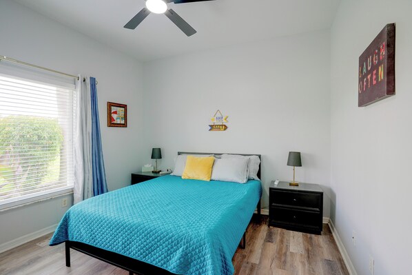 A bedroom with a bed, two bedside tables, a nightstand, a ceiling fan, a window with curtains, and a wall decoration.