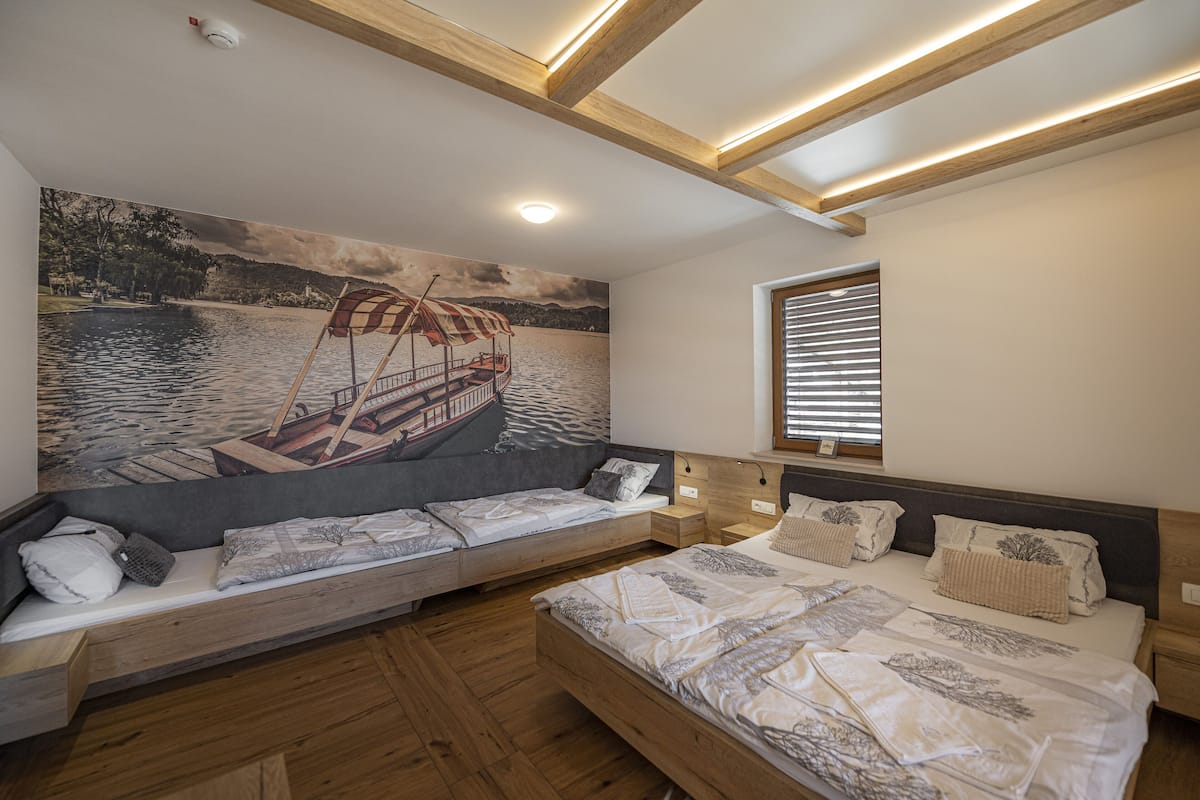 A bedroom with a large bed, a bench with cushions, a wooden floor, a painting of a boat on the wall, and a window with blinds.
