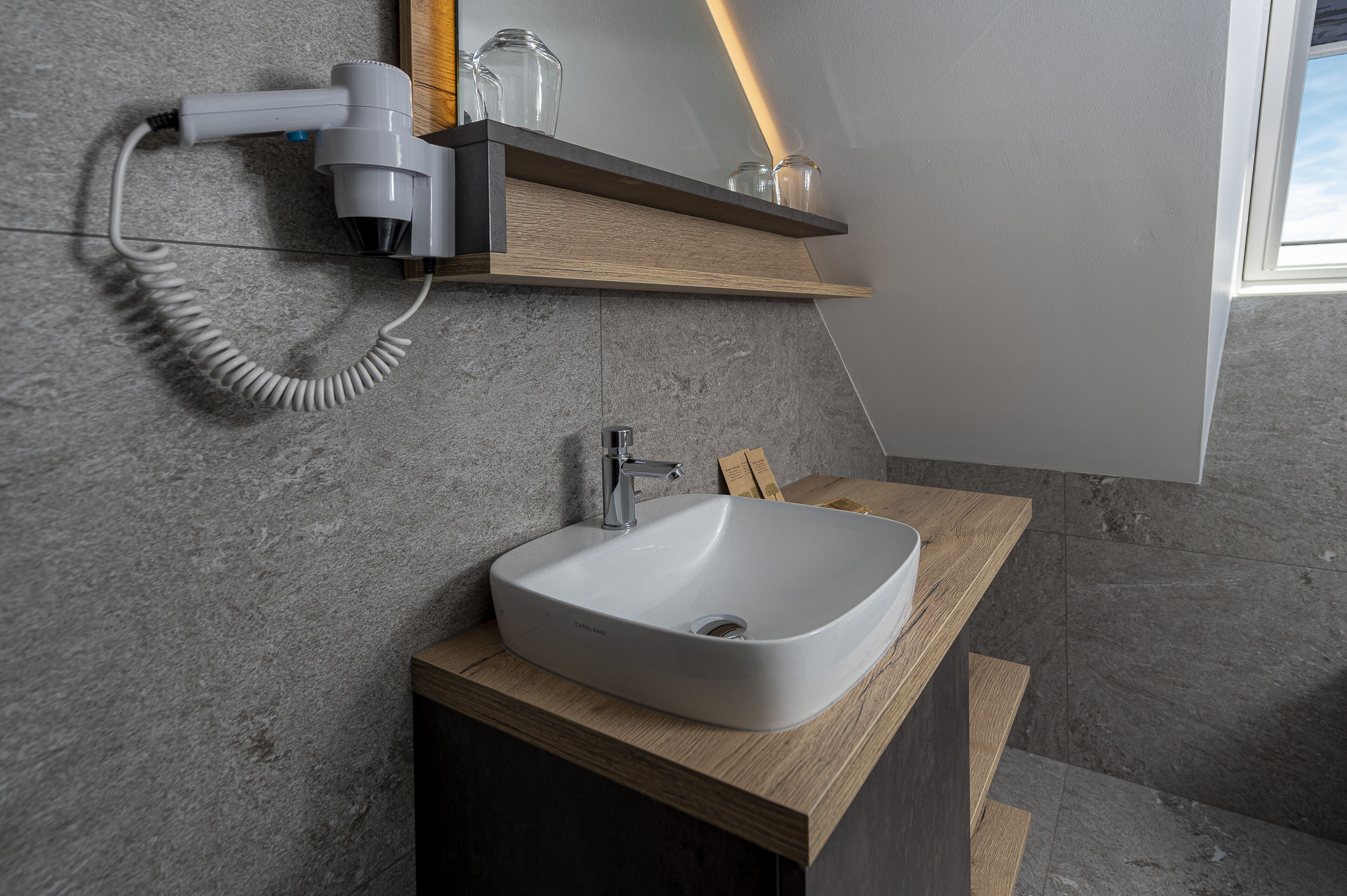 Een moderne badkamer met een witte wastafel, een aan de muur bevestigde haardroger en een houten plank boven de wastafel.