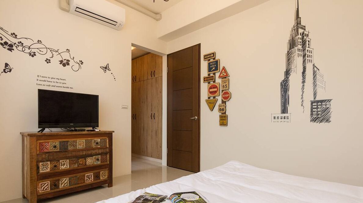 A bedroom with a bed, a TV on a wooden dresser, a wall with a cityscape mural, and a door leading to another room.