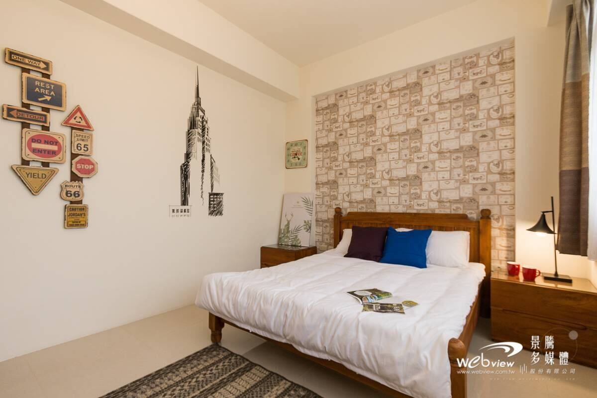 A bedroom with a bed, a nightstand, a wall with road signs, and a cityscape artwork.