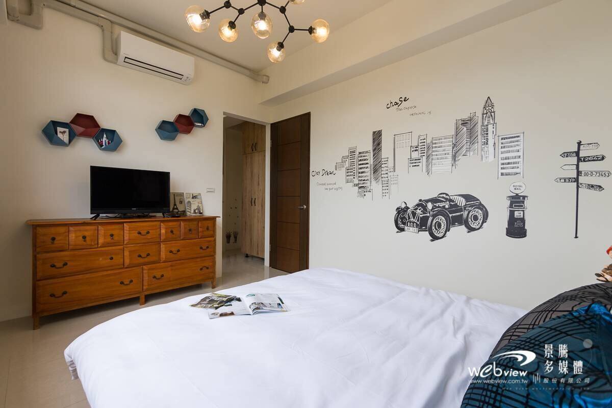 A bedroom with a bed, a wooden dresser, a flat-screen TV, and a wall with cityscape and car decals.