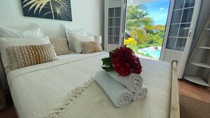 Une chambre avec un grand lit, du linge de lit blanc et une vue sur une piscine, visible à travers une porte coulissante ouverte.