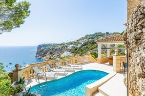 A swimming pool with lounge chairs, a stone wall, and a view of the sea and hillside houses. - Villa "Cala Llamp" with Panoramic Sea Views (PORT ANDRATX)