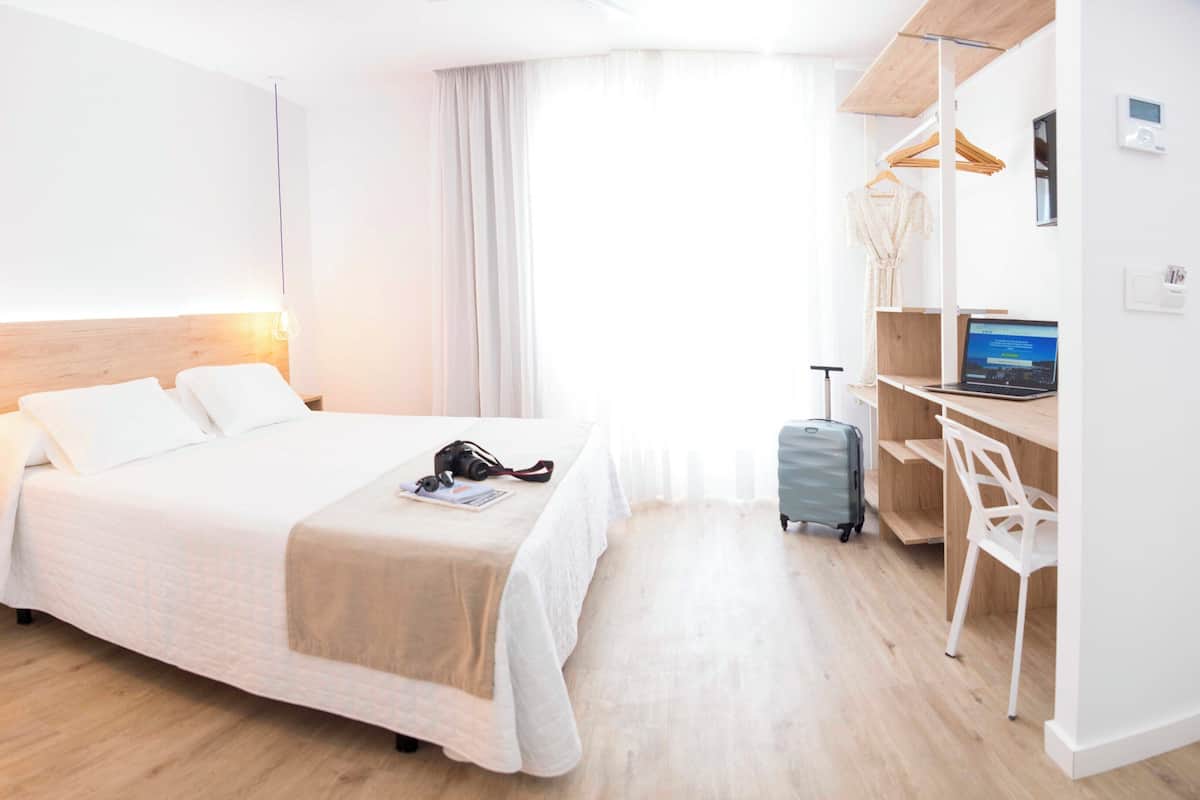 A modern hotel room with a large bed, a desk with a laptop, a suitcase, and a wardrobe.