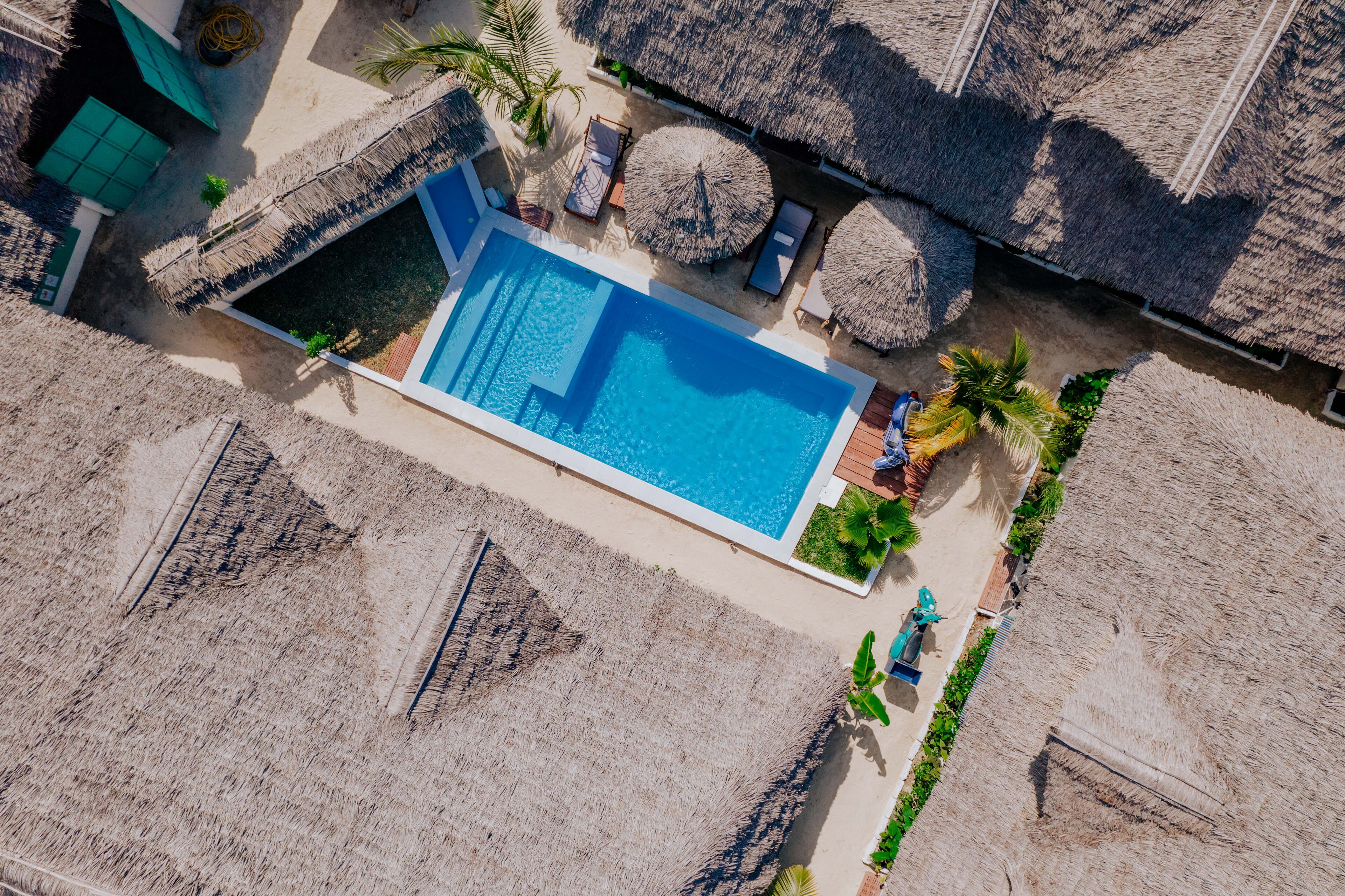 An aerial view of a resort with a rectangular swimming pool surrounded by thatched umbrellas and lounge chairs.