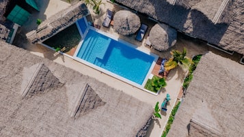 An aerial view of a resort with a rectangular swimming pool surrounded by thatched umbrellas and lounge chairs.